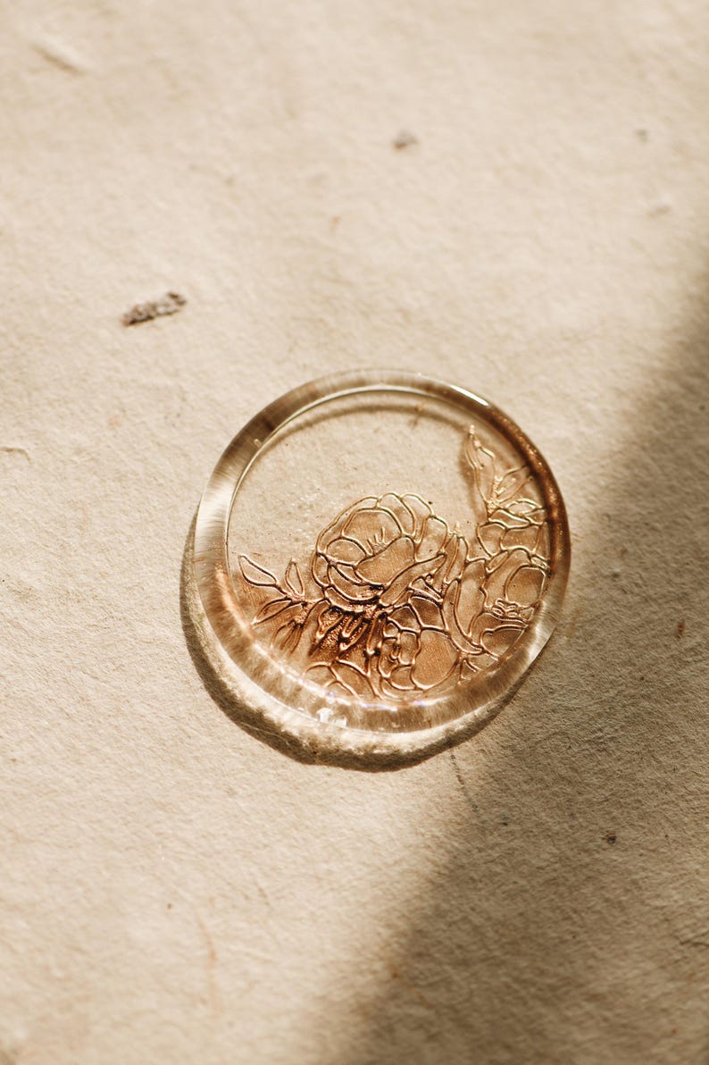 May include: A clear wax seal with a floral design in gold. The seal is round and has a raised edge.