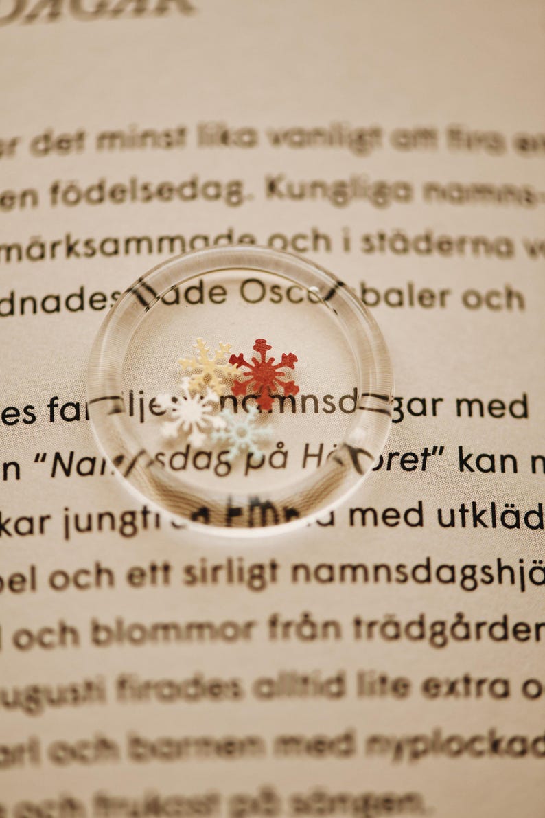 May include: A close-up of a magnifying glass with four small, decorative snowflakes inside. The snowflakes are red, white, and light blue. The magnifying glass is on a piece of paper with text in a foreign language.