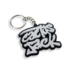 Könnte beinhalten: Schwarz-weißer Schlüsselanhänger mit dem Schriftzug "cactus jack" in einer stilisierten Schriftart. Der Schlüsselanhänger hat einen silbernen Schlüsselring und eine Kette. Der Text ist weiß, der Hintergrund ist schwarz.