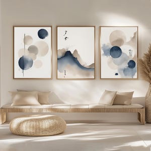 May include: Three framed watercolour art prints. Each print features abstract designs in shades of blue, beige, and grey. The artwork includes circles and mountain-like shapes, with some prints featuring Japanese calligraphy. The frames are a light brown colour.