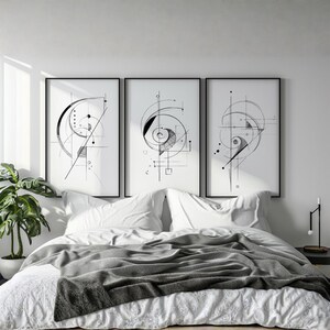 Könnte beinhalten: Drei gerahmte Kunstdrucke mit schwarzen geometrischen Designs auf weißem Hintergrund. Die Drucke werden über einem Bett mit weißer Bettwäsche und einer grauen Decke ausgestellt. Eine Topfpflanze ist links zu sehen.