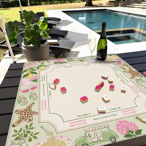 Puede incluir: Un tapete de mahjong cuadrado con centro color crema y borde decorativo con ilustraciones de limones, flores rosas, gafas de sol, tacones, alfombra de leopardo, pasta y un carrito de golf verde, con instrucciones de juego y fichas.