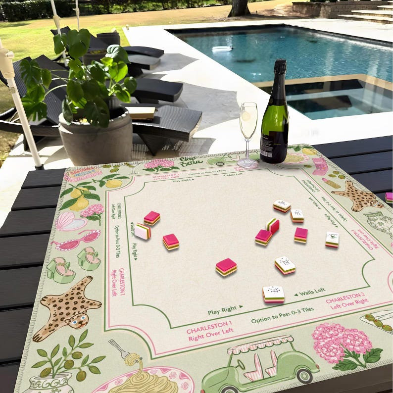 Puede incluir: Un tapete de mahjong cuadrado con centro color crema y borde decorativo con ilustraciones de limones, flores rosas, gafas de sol, tacones, alfombra de leopardo, pasta y un carrito de golf verde, con instrucciones de juego y fichas.