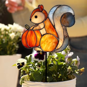 May include: A stained glass squirrel garden stake holding an orange pumpkin. The squirrel has a brown and orange body with a fluffy tail. The garden stake is placed in a white pot with colorful flowers.