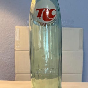 May include: A clear glass RC Cola bottle with a white and red logo. The bottle has a capacity of 10 fluid ounces. The bottle is empty and has a light green tint. The bottle is on a white surface.