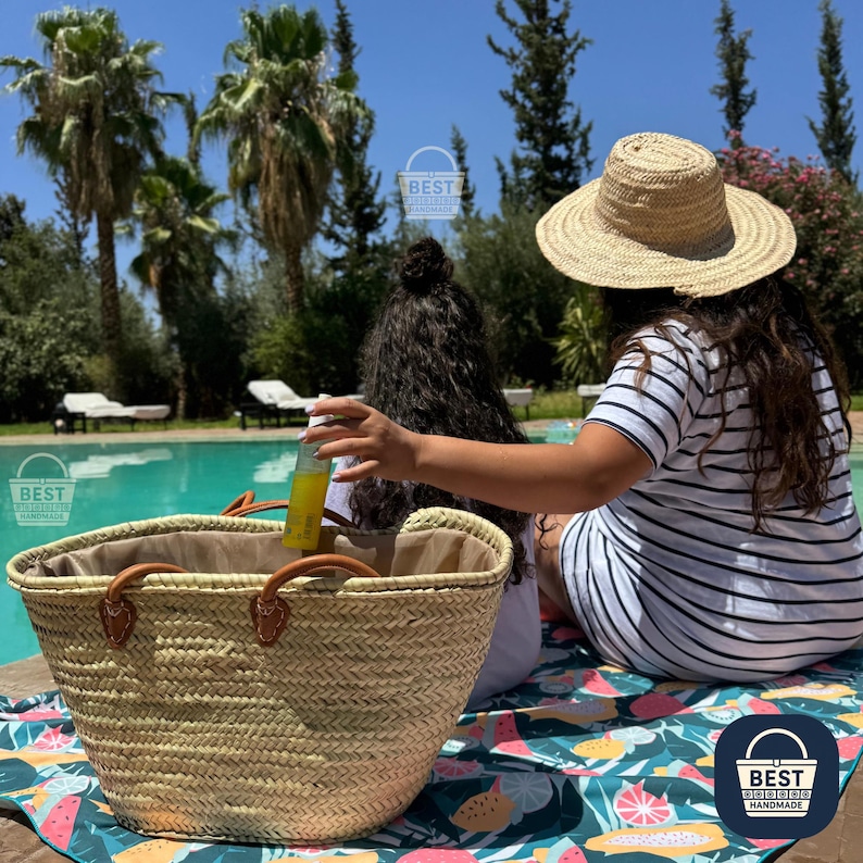 May include: A woven straw tote bag with brown leather handles sits near a pool. A person wearing a straw hat and a striped dress is seated nearby. The scene is outdoors on a sunny day.