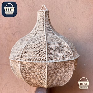 Può includere: Lampada a sospensione beige intrecciata, dalla forma bulbosa. La trama aperta è visibile su tutta la superficie. Ha un anello per appenderla. Le parole "BEST HANDMADE" sono stampate su un quadrato blu.