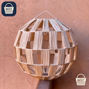 Puede incluir: Lámpara colgante esférica de fibras naturales tejidas. El diseño de tejido abierto permite el paso de la luz. El texto "BEST HANDMADE" es visible en la parte superior izquierda e inferior derecha. La lámpara se cuelga de un lazo.