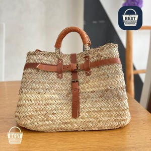 Puede incluir: Bolso tote de paja tejida con asas y correas de cuero marrón. El bolso tiene forma rectangular con un patrón tejido y cierre de hebilla. Las asas están envueltas en cuero marrón. Hecho a mano.