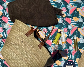 May include: A woven straw tote bag with brown leather handles sits on a colorful picnic blanket with a fruit pattern. A dark brown towel and several bottles of sunscreen and lotion are also on the blanket.