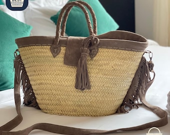 Puede incluir: Bolso tote de paja tejida con detalles de ante marrón, que incluye un cierre de solapa, asas trenzadas y una correa larga para el hombro. El bolso presenta flecos en los laterales y una borla decorativa.