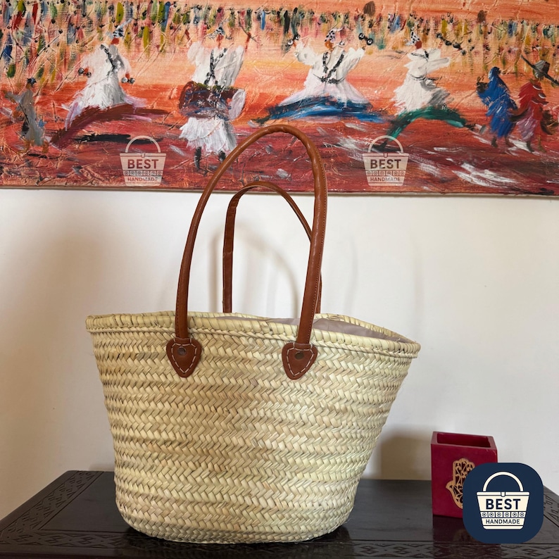 May include: A woven straw tote bag with brown leather handles and accents. The bag has a natural, light beige color and a spacious interior. The handles are attached with brown leather patches. The bag is sitting on a dark surface.