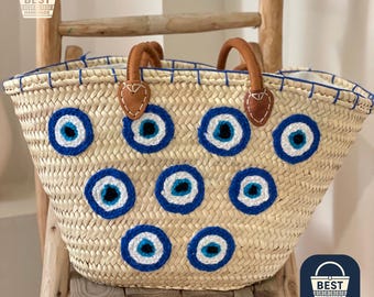 May include: Woven straw tote bag with brown leather handles and blue and white evil eye designs. The bag has a rectangular shape and is decorated with multiple embroidered eye motifs. The bag is handmade.