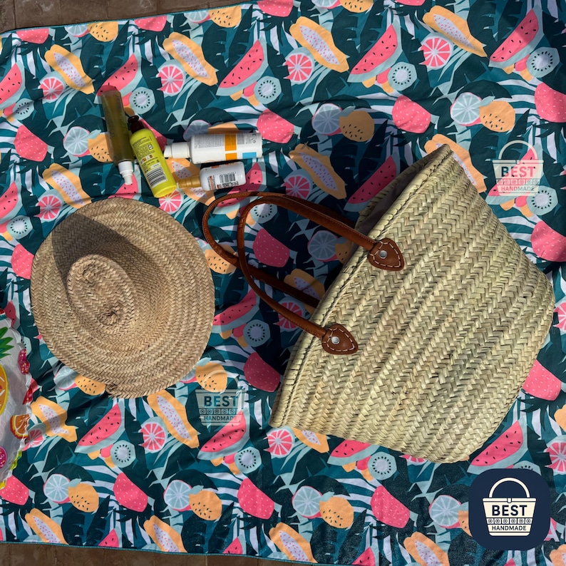 May include: A woven straw tote bag with brown leather handles sits on a colorful beach blanket. The blanket features a tropical fruit pattern in pink, yellow, and green. A straw hat and sunscreen bottles are also visible.