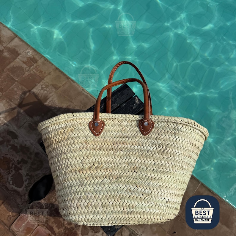 May include: A woven straw tote bag with brown leather handles and accents. The bag is sitting on a wooden surface next to a pool of turquoise water. The bag has a rectangular shape and is designed for carrying items.