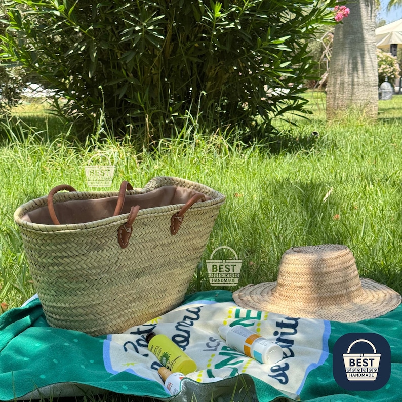 May include: A woven straw tote bag with brown leather handles and a matching straw sun hat sit on a green towel. The towel has a blue and white design with bottles of sunscreen and other beach essentials.