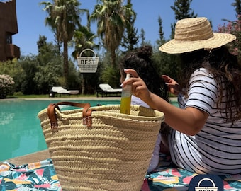 May include: A woven straw tote bag with brown leather handles sits on a colorful patterned towel. The bag contains a bottle of liquid. A person wearing a straw hat and striped shirt is near the bag. The scene is outdoors near a pool.