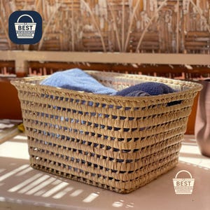 May include: A rectangular woven basket made of natural fibers, filled with blue towels. The basket has a textured, open weave design and is labeled "BEST HANDMADE".