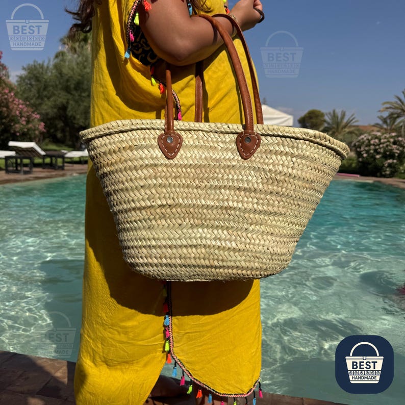 May include: A woven straw tote bag with brown leather handles and accents. The bag is held by a person wearing a yellow dress. The background includes a swimming pool and palm trees. The bag has a natural, light brown color.