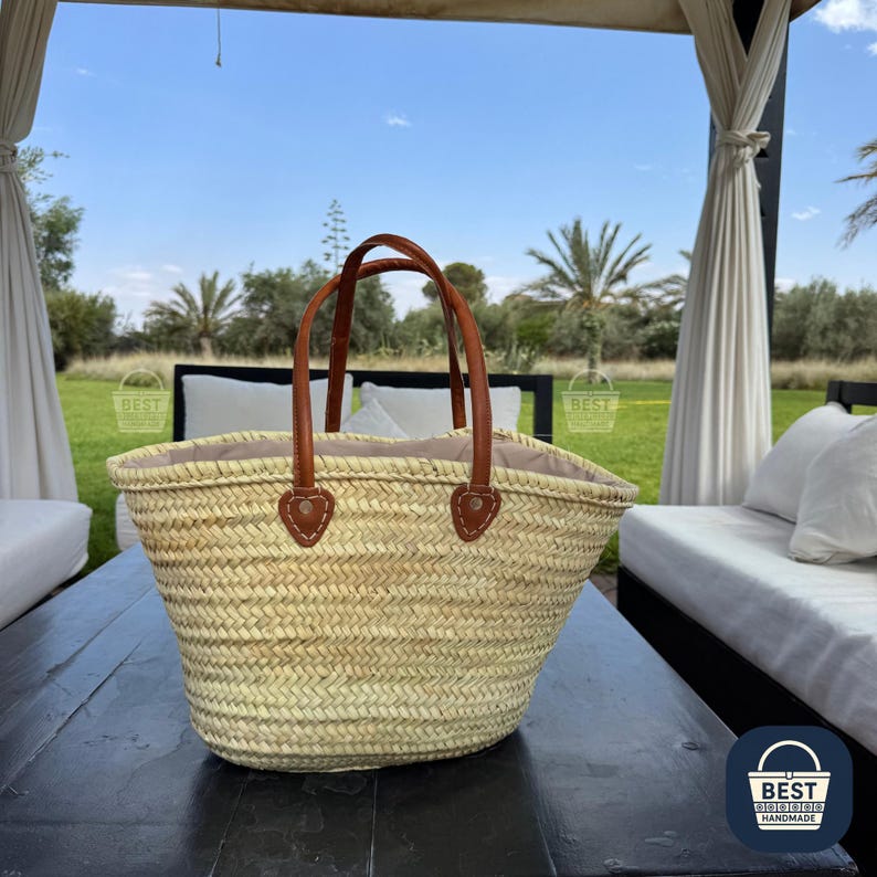 May include: A woven straw basket with brown leather handles and accents. The basket has a light-colored interior and is sitting on a dark wooden surface. The handles are attached with brown leather patches.