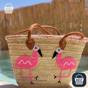 May include: Woven straw tote bag featuring two embroidered pink flamingo designs. It has brown leather handles and pink stitching. The bag is labelled "BEST HANDMADE".