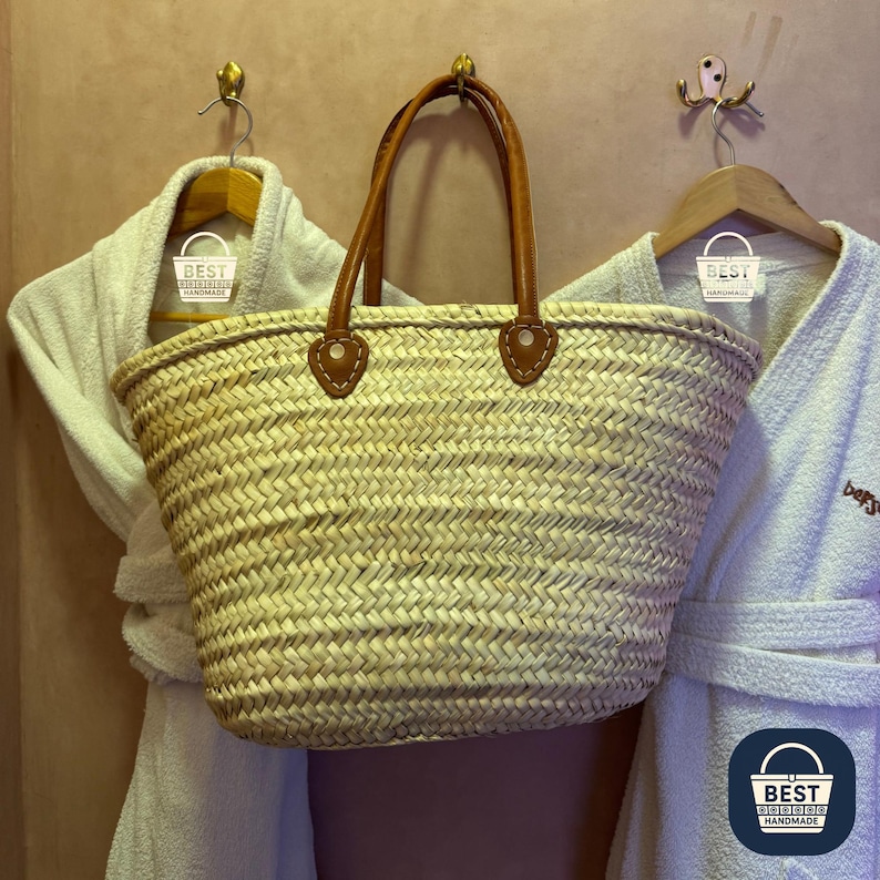 May include: Woven straw tote bag with brown leather handles and accents. The bag is rectangular with a rounded top and is hanging on a wall. Two white bathrobes hang on either side of the bag.