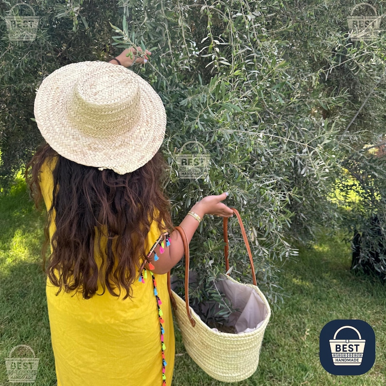 May include: A straw hat with a wide brim and a woven basket with a brown leather handle. The person is wearing a yellow dress and is reaching towards a tree with green leaves. The basket has a white lining.