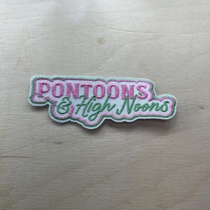 Könnte beinhalten: Gestickter Aufnäher mit den Worten "Pontoons & High Noons" in rosa und grüner Schrift. Der Aufnäher hat einen weißen Rand und liegt auf einem hellen Holzmaserungshintergrund. Der Aufnäher ist wahrscheinlich für Kleidung oder Accessoires.