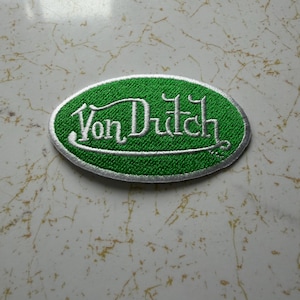 Pode incluir: Patch bordado com o texto "Von Dutch" em escrita branca sobre um fundo oval verde. O patch tem uma borda prateada, ideal para roupas ou acessórios.