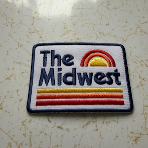May include: Embroidered patch with the words "The Midwest" in navy blue, above a rainbow design in red, orange, and yellow. Below the text are three horizontal stripes in yellow, orange, and red. The patch has a white background and a navy blue border.