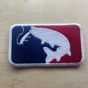 May include: Embroidered patch featuring a stylized baseball player silhouette. The patch has a rectangular shape with a white border. The design incorporates blue, white, and red colors, with a baseball player in white.