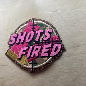 May include: Embroidered patch with the words "SHOTS FIRED" in pink glitter lettering. The patch features a pink target design over a camouflage background, outlined with gold glitter.