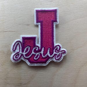 以下が含まれることがあります： ピンク色の刺繍パッチ。文字「J」の形をしており、筆記体で「Jesus」と書かれています。パッチは白い縁取りがあり、明るい木目調の背景に配置されています。