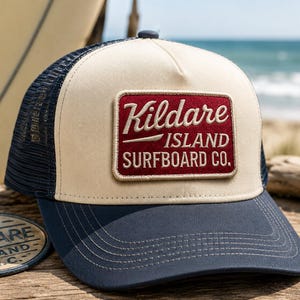 May include: A trucker hat with a cream-colored front panel, navy blue mesh back, and a navy blue brim. A rectangular patch on the front features a maroon background with white embroidered text that reads Kildare ISLAND SURFBOARD CO.