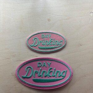 May include: Two embroidered patches with the words "DAY Drinking" in a script font. The patches are oval-shaped with a pink background and a green border and lettering. The patches are on a light wood surface.
