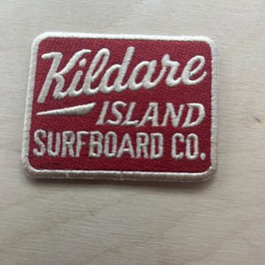 May include: A rectangular red patch with white embroidered text. The patch reads "Kildare ISLAND SURFBOARD CO." in a stylized font. The patch has a white border and is set against a light wood grain background.