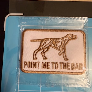 May include: A rectangular embroidered patch with a white background and a brown border. It features a brown outline of a dog and the text "POINT ME TO THE BAR" in brown thread. The patch is placed on a blue cutting mat with inch markings.