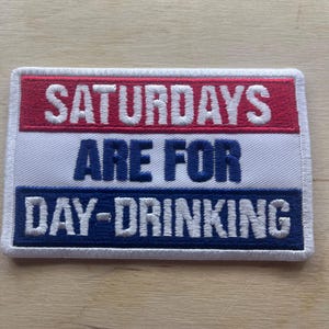 May include: Embroidered patch with the text "SATURDAYS ARE FOR DAY-DRINKING" in white, blue, and red. The patch has a rectangular shape with a white border. The text is in a bold, sans-serif font.