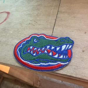 May include: An embroidered patch featuring a green alligator head with sharp white teeth, set against a circular background of blue and red. The design is outlined in blue thread, showcasing a detailed profile of the reptile with a focused expression.