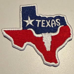 May include: An embroidered patch in the shape of the state of Texas. The top section is dark blue with a white star and the word TEXAS in white lettering. The bottom section is red, featuring a white longhorn skull silhouette in the centre.