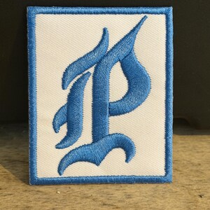 Pode incluir: Um emblema retangular bordado com fundo branco e uma borda azul espessa. Centralizada no emblema está uma letra P grande e estilizada em uma fonte azul ousada de inspiração gótica com curvas fluidas e pontas afiadas.