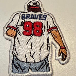 May include: An embroidered patch depicting a figure from the back wearing a white baseball jersey with the word BRAVES and the number 98 in red. The figure wears a white cap with a W, blue jeans, and has tan skin tones with visible arm details.