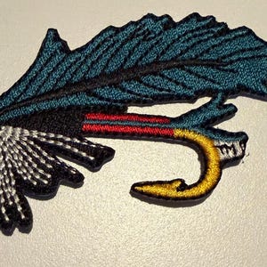May include: An embroidered patch depicting a fishing fly lure. The design features a large teal feather-like element, a black base, white accent stitching, a horizontal red and blue striped section, and a curved yellow hook shape at the bottom.