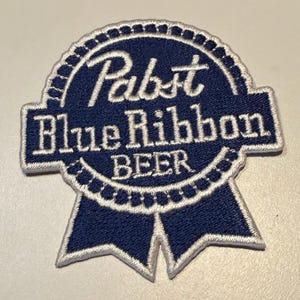 May include: A circular embroidered patch featuring a dark blue background with white stitched borders and text. The design includes the words Pabst Blue Ribbon BEER in white lettering, shaped like a ribbon award with two tails at the bottom.