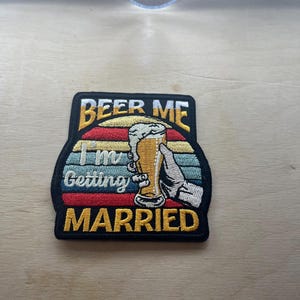 Może przedstawiać: Haftowana naszywka z napisem "BEER ME I'm Getting MARRIED". Naszywka przedstawia dłoń trzymającą kufel piwa z pianą, na tle w paski w kolorach czerwonym, żółtym i niebieskim.