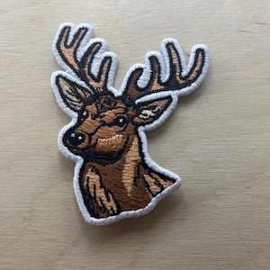 May include: Embroidered patch featuring a detailed brown deer head with large antlers. The patch has a white border and is designed with intricate stitching, showcasing the deer's features. Ideal for jackets, bags, or other fabric items.