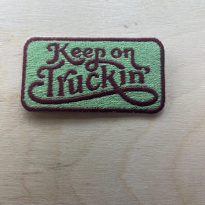 Könnte beinhalten: Gestickter Aufnäher mit dem Schriftzug "Keep on Truckin'" in bordeauxfarbener Schrift auf hellgrünem Hintergrund. Der Aufnäher hat einen bordeauxfarbenen Rand und eine rechteckige Form. Der Aufnäher liegt auf einer hellen Holzoberfläche.