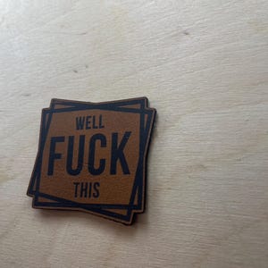 May include: A brown leather patch with the words "WELL FUCK THIS" in black block letters. The patch has a layered square design with black borders. The background is a light wood grain.