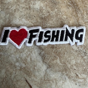May include: An embroidered patch featuring the text I, a red heart symbol, and the word FISHING. The letters are black with a thick white border, and the heart is red with a white border, creating a high-contrast design for fabric application.