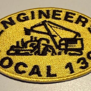 May include: An oval-shaped embroidered patch with a yellow background and a black border. The patch features a black embroidered graphic of a construction crane and the text "ENGINEERS" at the top and "LOCAL 139" at the bottom in black thread.